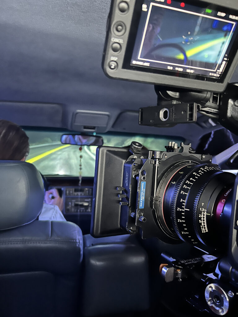 A cinema camera sits in the backseat of a car. A roadway with double yellow lines is projected through the windshield.